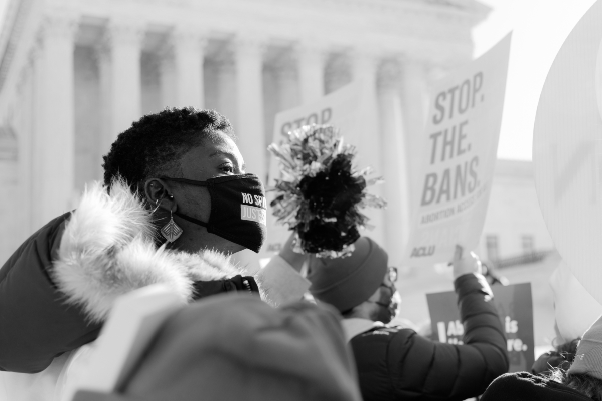 Black woman in a mask looking powerful in front of the courthouse next to a reproductive rights sign that reads 'Stop the Bans'